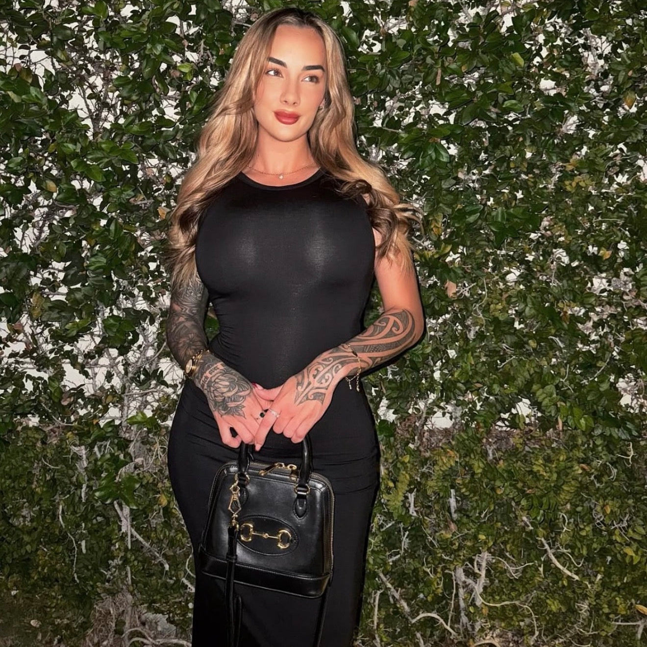 Woman in a black outfit with tattoos holding a black handbag against a green leafy background