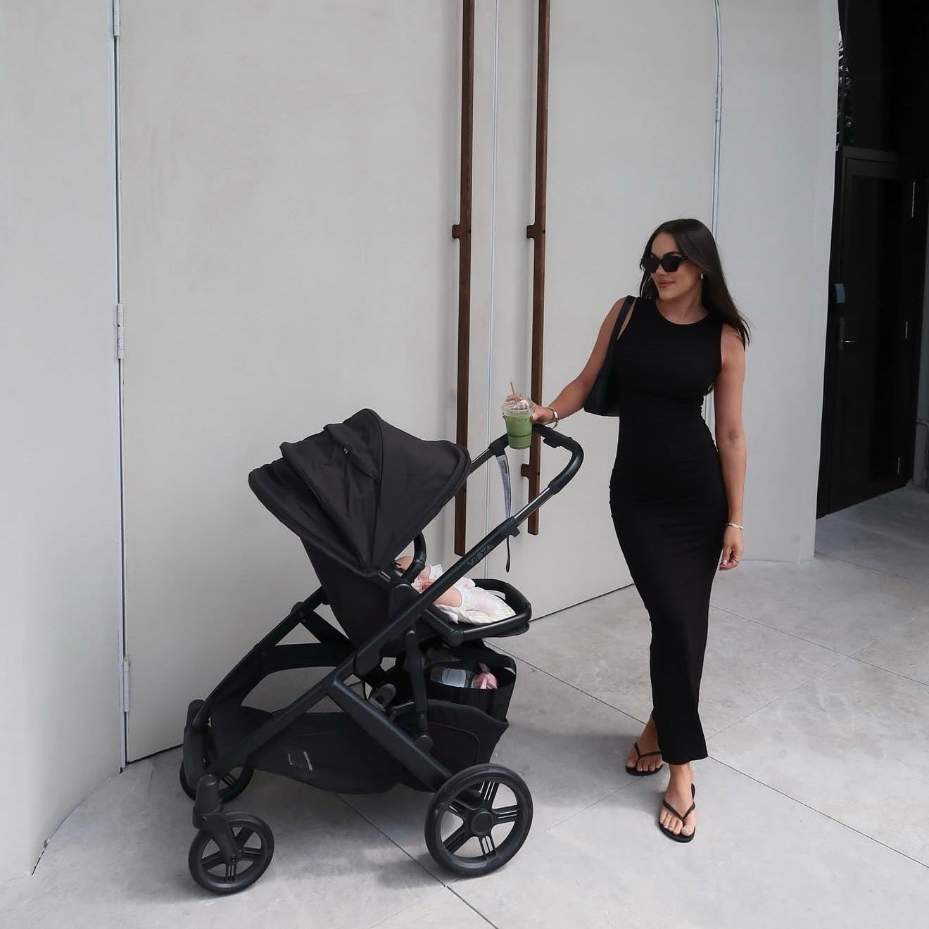 Woman in black outfit pushing a black stroller against a white wall.