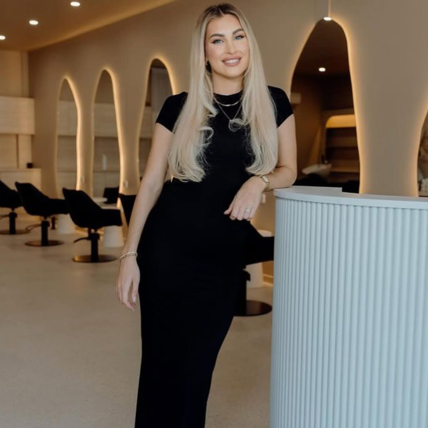 Woman in a black outfit standing in a modern interior setting with arches and chairs.