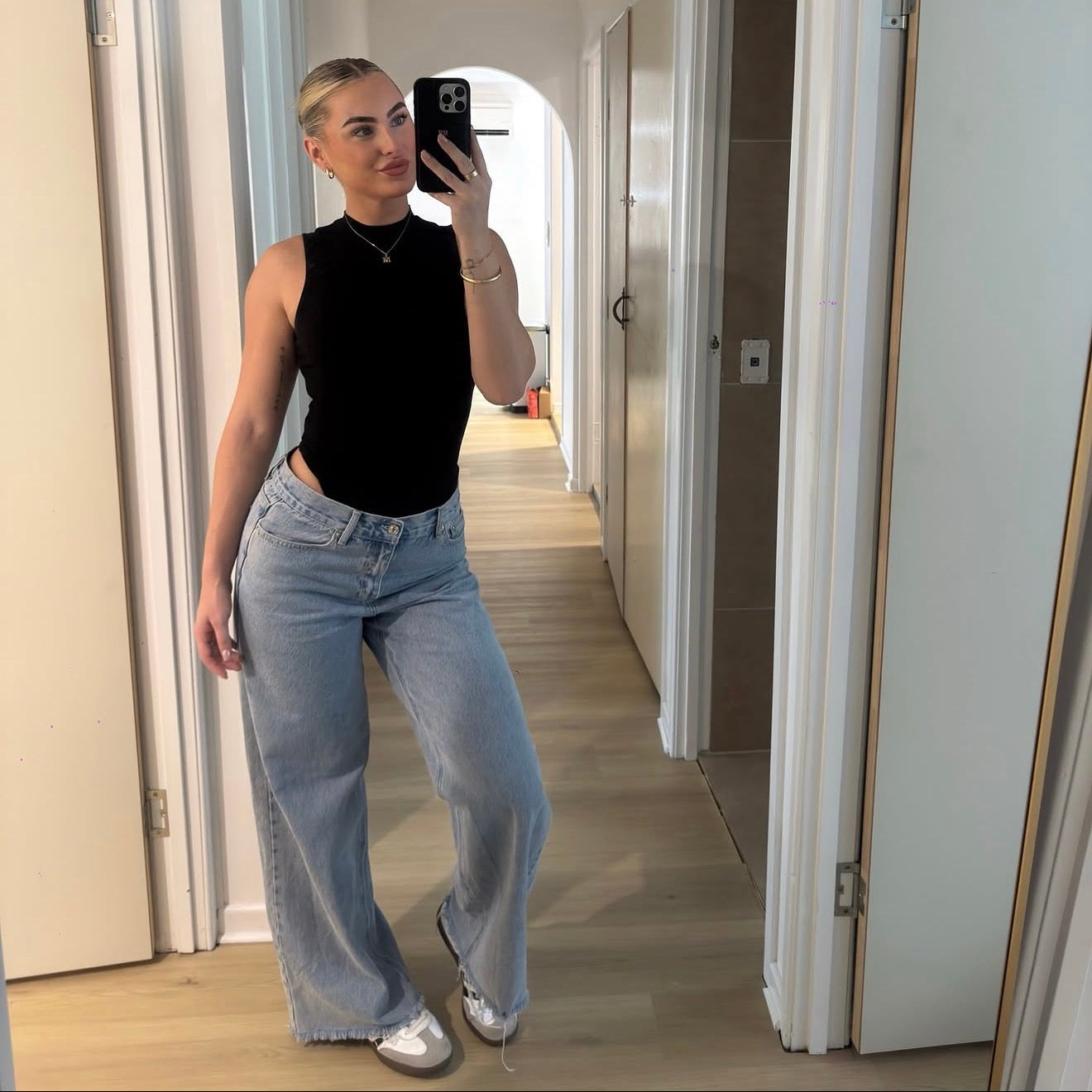 Person taking a mirror selfie wearing a black sleeveless top and light blue jeans in a hallway.