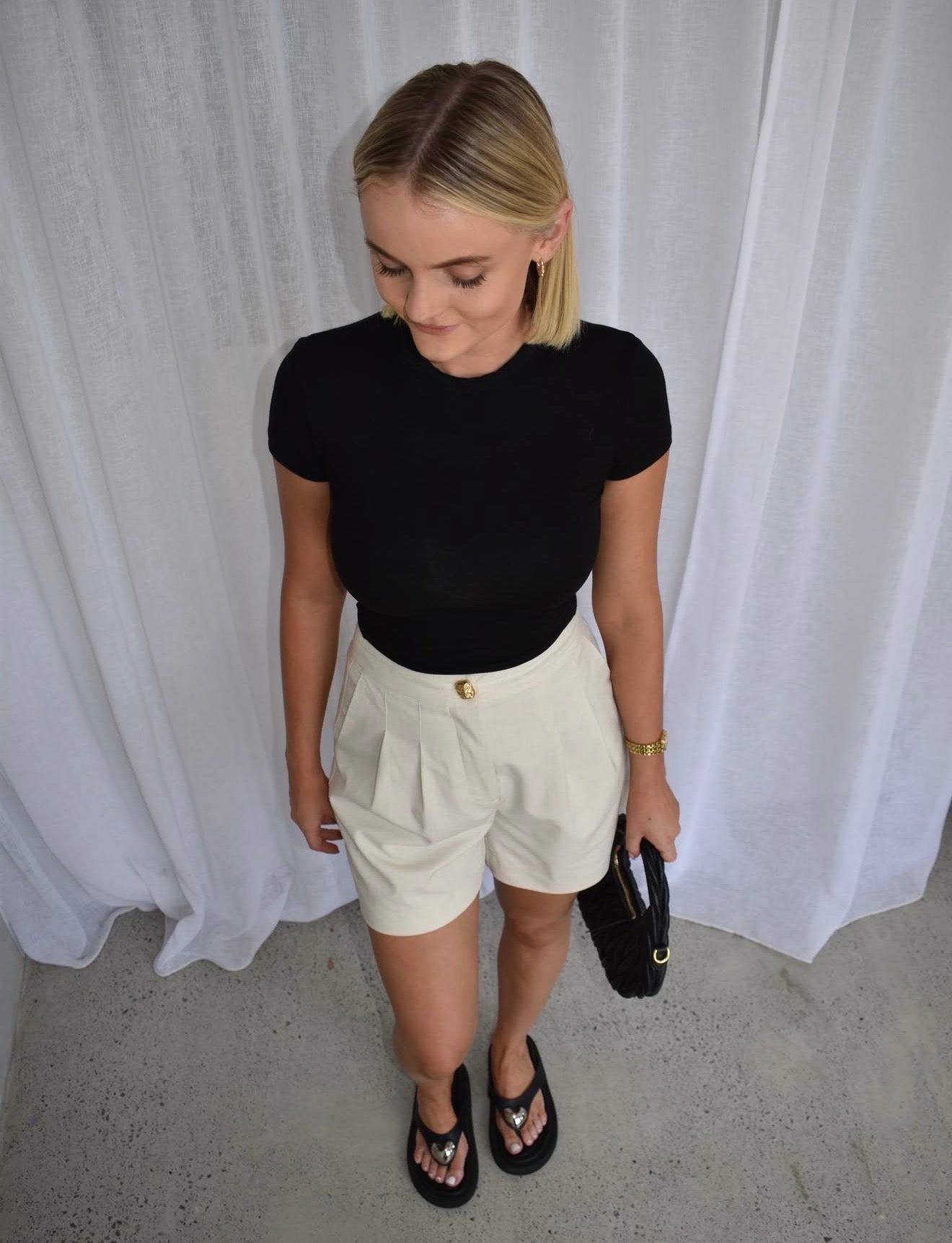 Woman wearing a black top and white shorts against a white curtain background