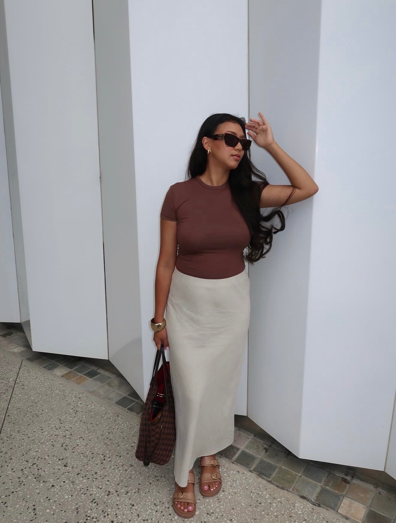 Woman in a brown top and white skirt standing against a white wall.