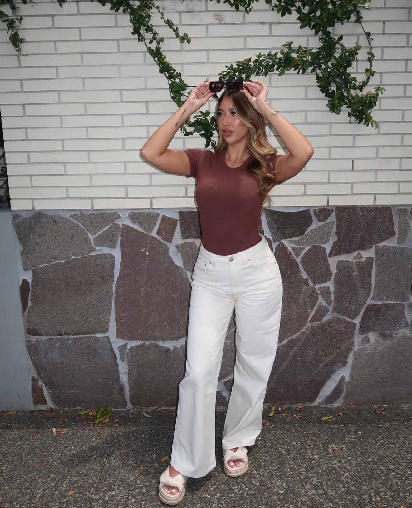 Woman in a brown top and white pants standing against a brick wall with stone base.