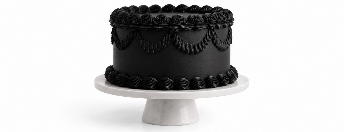 A black frosted cake with decorative piped borders sits on a white pedestal stand against a white background, with the text 'SULE turns 3' above it.