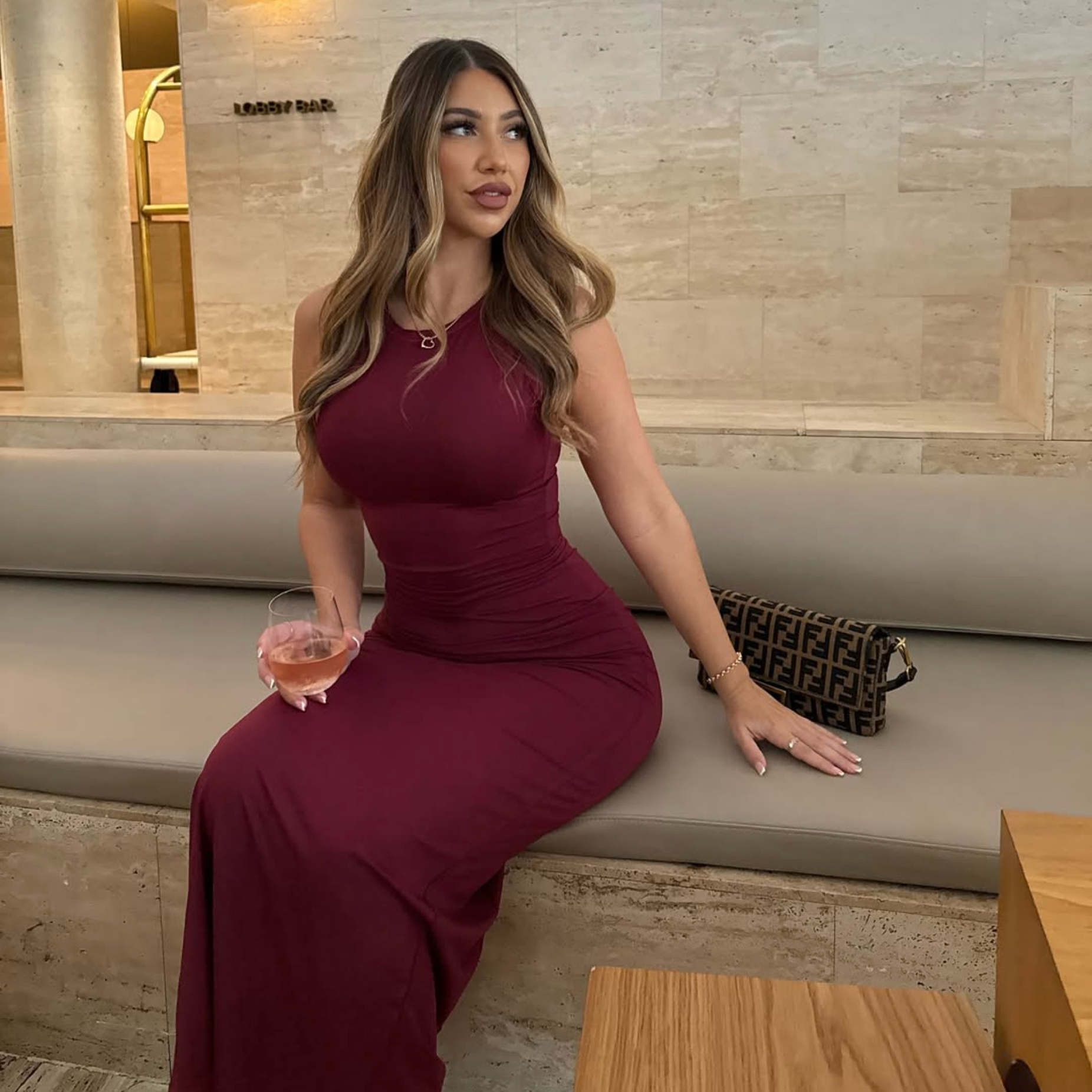 Woman in a burgundy dress sitting on a bench holding a glass, with a Fendi handbag beside her.