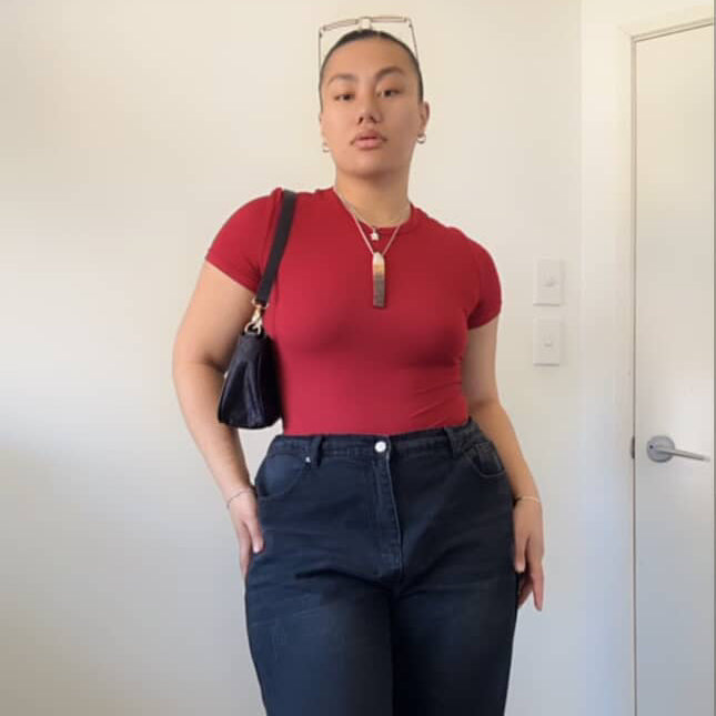 Person wearing a red top and dark jeans indoors against a white wall.