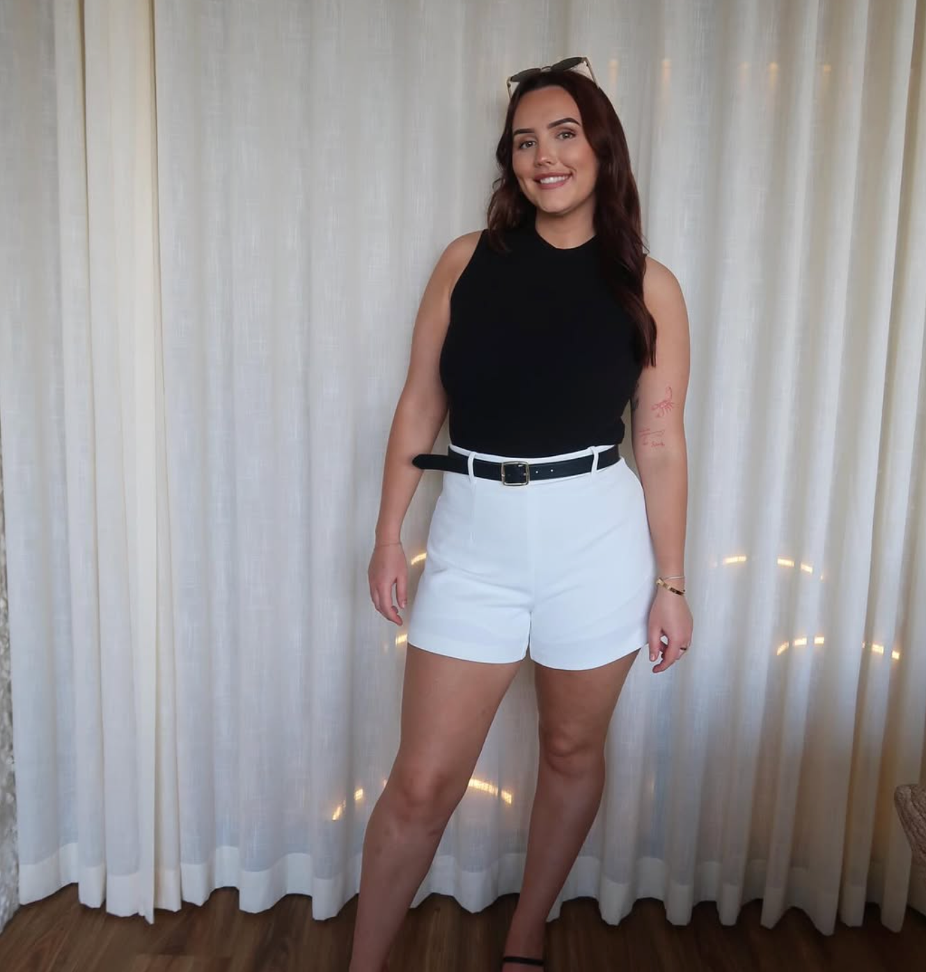 Woman wearing a black sleeveless top and white shorts standing in front of white curtains.